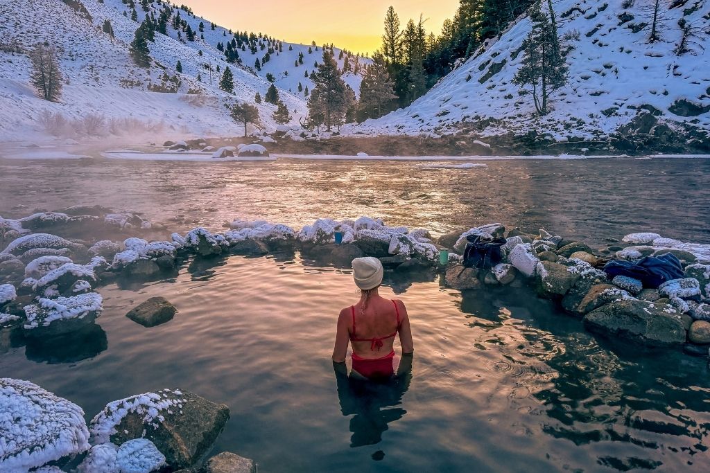 Kate in a red swimsuit and white beanie soaks in the steaming waters of Cove Hot Springs, surrounded by snow-covered rocks, as the golden glow of sunrise reflects off the river and the snowy hills in the background.