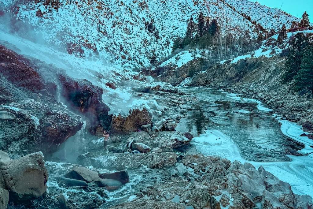 Steaming waters flow over rocky terrain into the icy river at Kirkham Hot Springs near Stanley, Idaho, with snow-covered hills and evergreen trees lining the landscape under a clear blue sky.