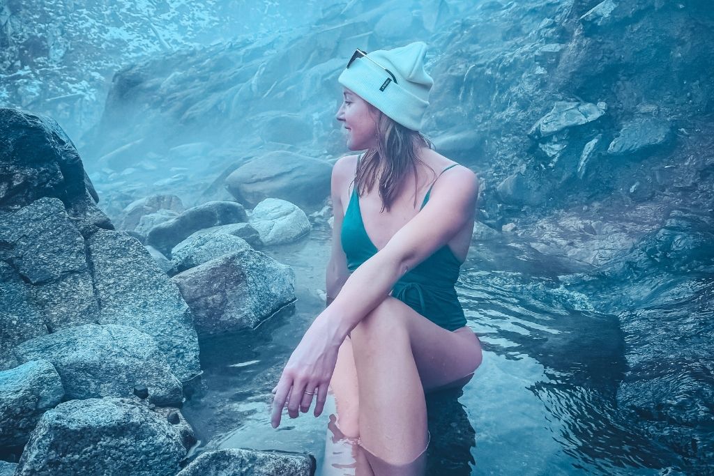 Kate, in a green swimsuit and white beanie, sits in the warm waters of Pine Flats Hot Springs, surrounded by misty steam and rugged, rocky terrain, with a serene expression on her face.