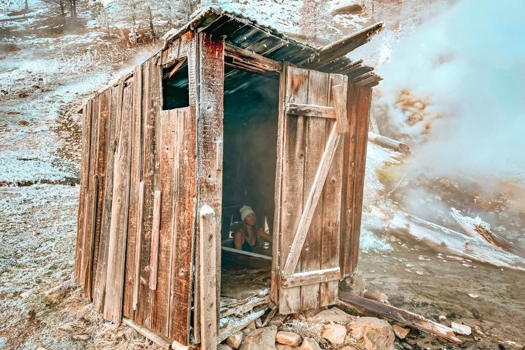 A rustic, weathered wooden hut stands at Bonneville Hot Springs, with steam rising from the nearby geothermal water, while Kate in a white beanie soaks in a small tub inside the open doorway.