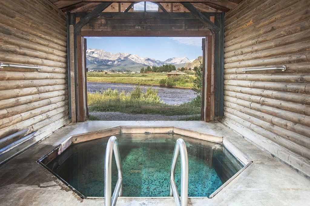 A serene indoor hot spring pool at Mountain Village Hot Springs, framed by a rustic log cabin door, opens to a stunning view of a flowing river and rugged mountain peaks under a clear blue sky.