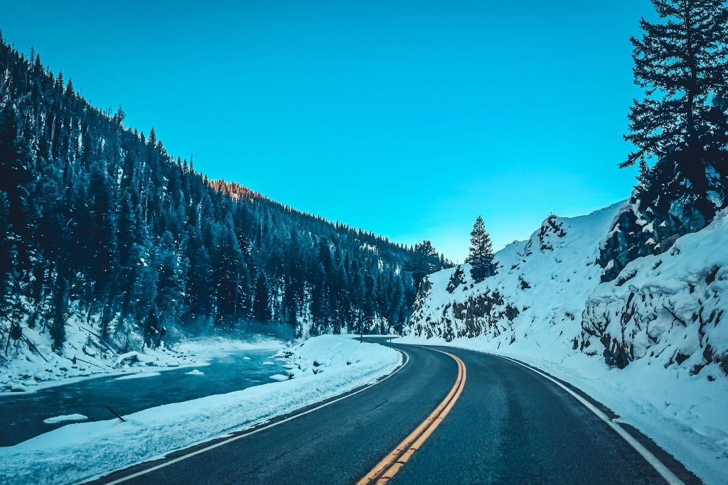 A winding mountain road curves alongside a partially frozen Salmon River, surrounded by snow-covered trees and rocky slopes under a clear blue sky.