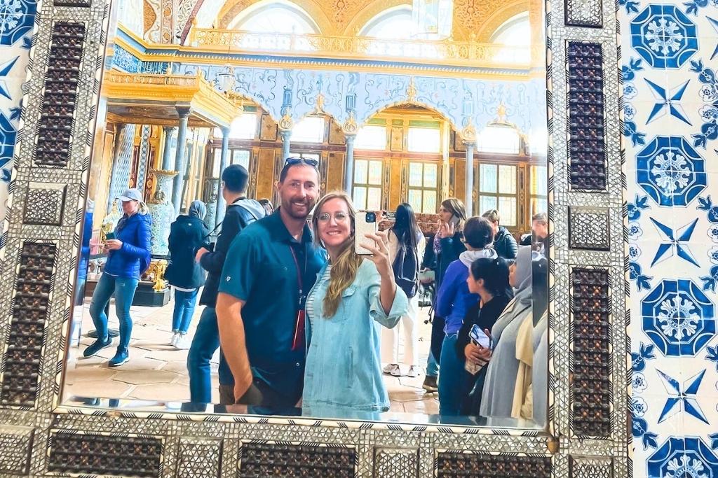 Kate and her husband take a mirror selfie inside Topkapi Palace, surrounded by intricate blue and white tilework, with a golden, ornately decorated interior and other visitors exploring the historic site.