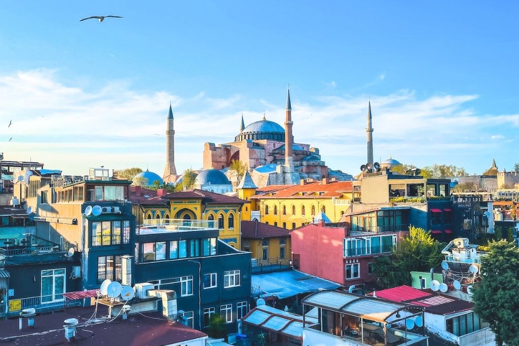 A scenic view of Istanbul's colorful rooftops with the iconic Hagia Sophia and its towering minarets in the background under a bright blue sky.