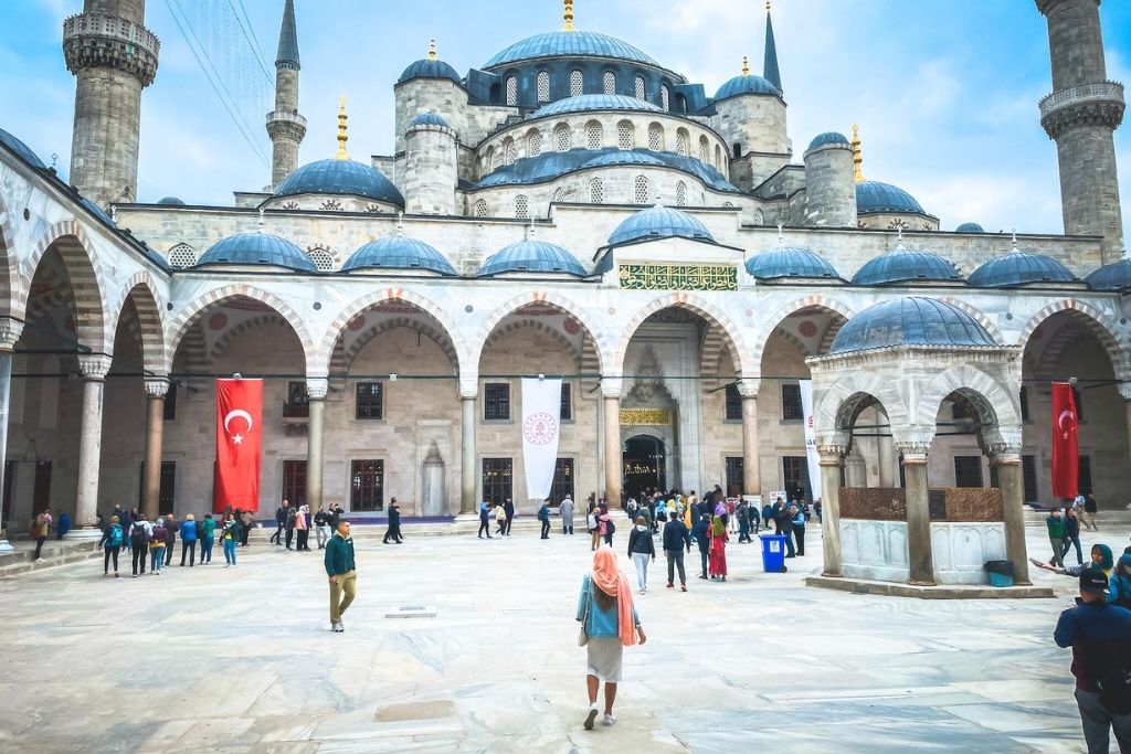 The courtyard of the Blue Mosque in Istanbul, Turkey, featuring grand domes, arched walkways, and Turkish flags, with visitors exploring the historic site under a cloudy sky.