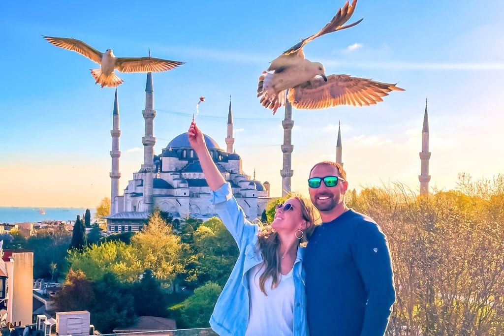 A smiling Kate and her husband poses on a rooftop in Istanbul with the Blue Mosque in the background, as Kate playfully feeds seagulls soaring above them at sunset.