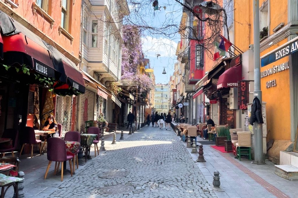 A charming cobblestone street in Istanbul’s Kadıköy neighborhood, lined with colorful buildings, cozy cafés with outdoor seating, and pedestrians strolling under hanging lights and blooming wisteria.