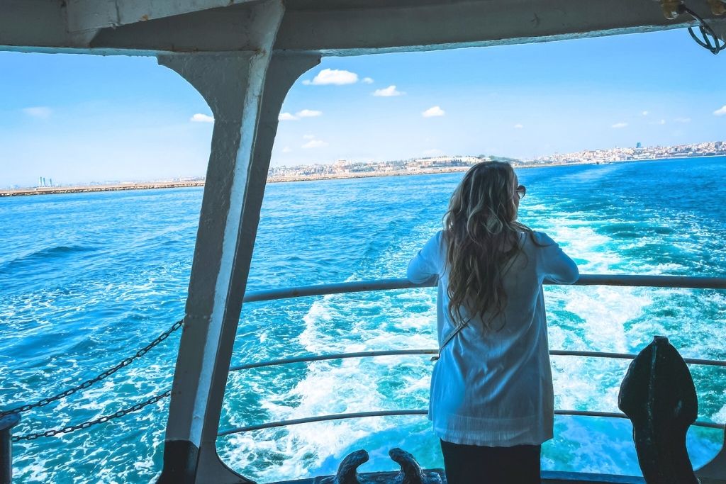 Kate, with long wavy blonde hair, wearing sunglasses and a light-colored cardigan, stands at the back of a ferry gazing at the deep blue waters of the Bosphorus with Istanbul’s skyline in the distance.