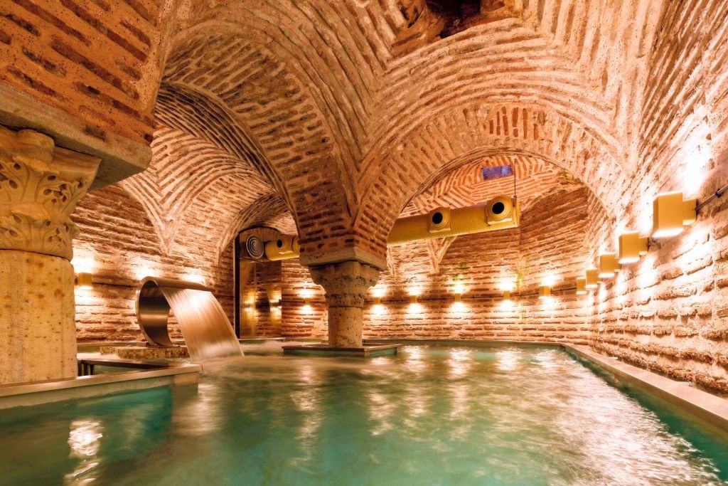 A luxurious indoor pool with cascading water features, set within the historic brick-arched interior of the Hagia Sofia Mansions Istanbul, Curio Collection by Hilton, illuminated by warm lighting.