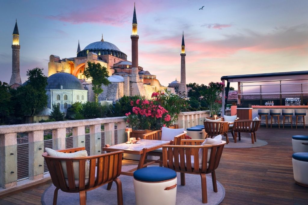 A rooftop terrace at the Four Seasons Hotel Istanbul at Sultanahmet, featuring elegant wooden seating, blooming flowers, and a breathtaking view of the illuminated Hagia Sophia at sunset.