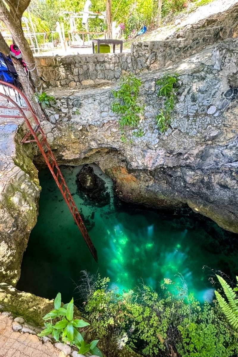 A natural limestone sinkhole in Jamaica glows with crystal-clear turquoise water, accessed by a steep red ladder and surrounded by lush greenery and stone walls.