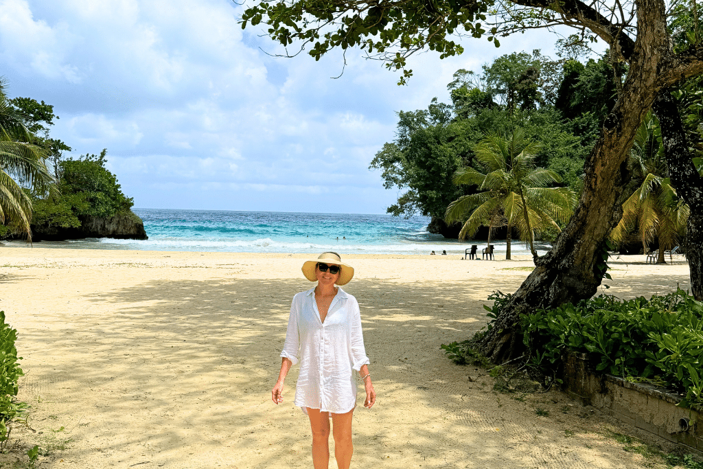 Kate stands on a quiet sandy beach in Jamaica, wearing a wide-brimmed sunhat and white cover-up, with turquoise waves and lush palm trees in the background.