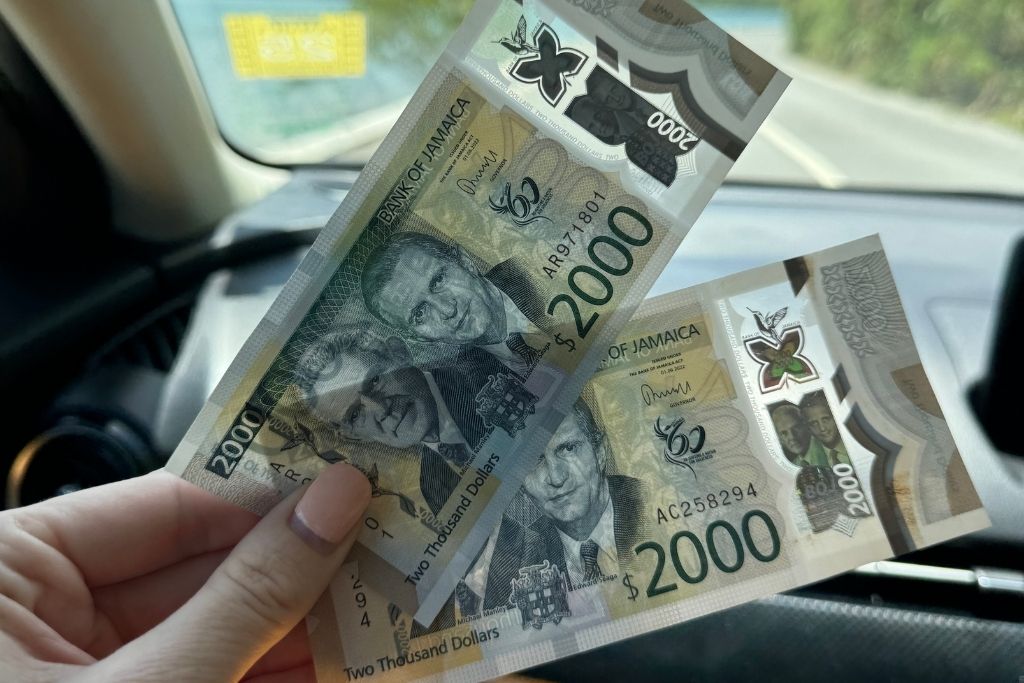 A hand holds two Jamaican $2000 banknotes inside a car, showing portraits of national leaders and crisp currency design in natural daylight.