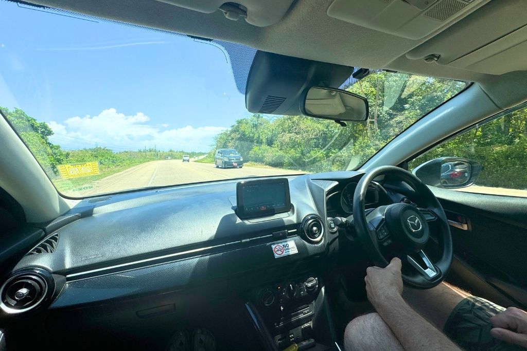 View from inside a car driving on a sunny Jamaican highway, with a clear blue sky, green roadside vegetation, and a right-hand steering wheel visible.