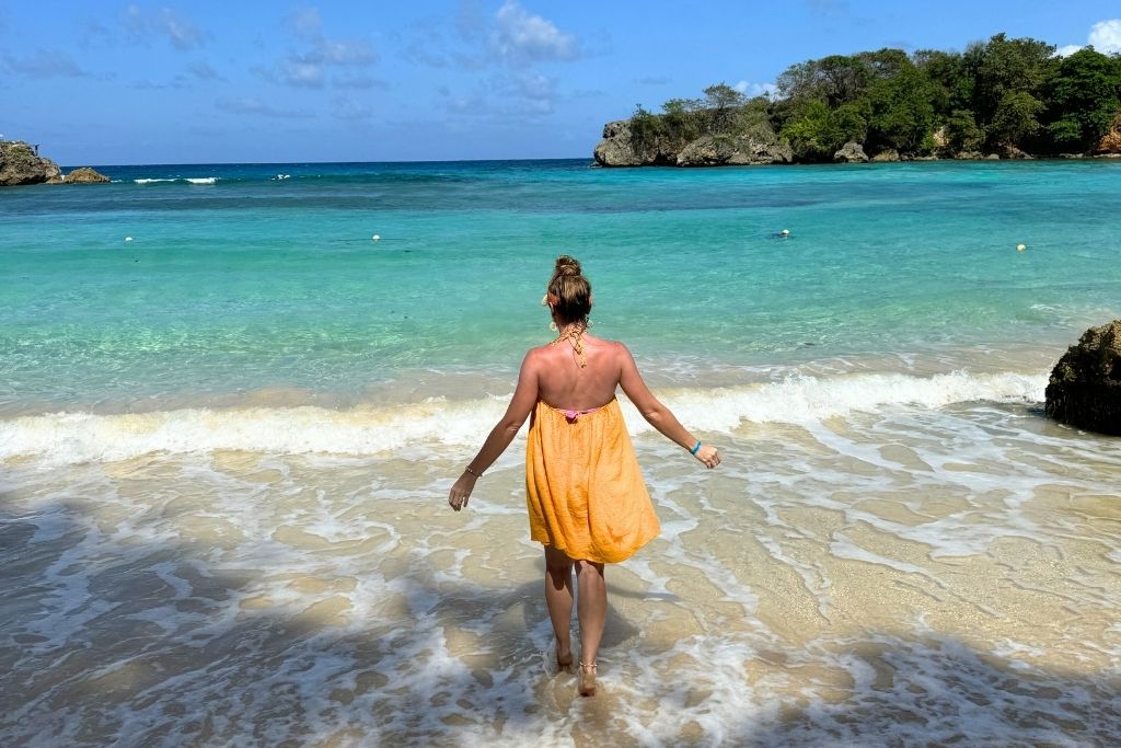 Kate walks barefoot into the gentle waves on a secluded Jamaican beach, wearing a yellow sundress with turquoise water and rocky cliffs in the distance.