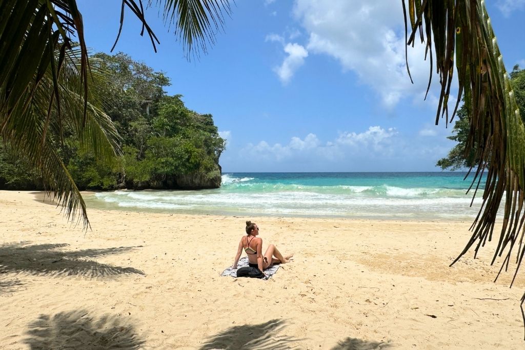 Kate relaxes on a towel under the shade of palm trees at a quiet Jamaican beach, gazing out at turquoise waves and lush cliffs under a sunny sky.