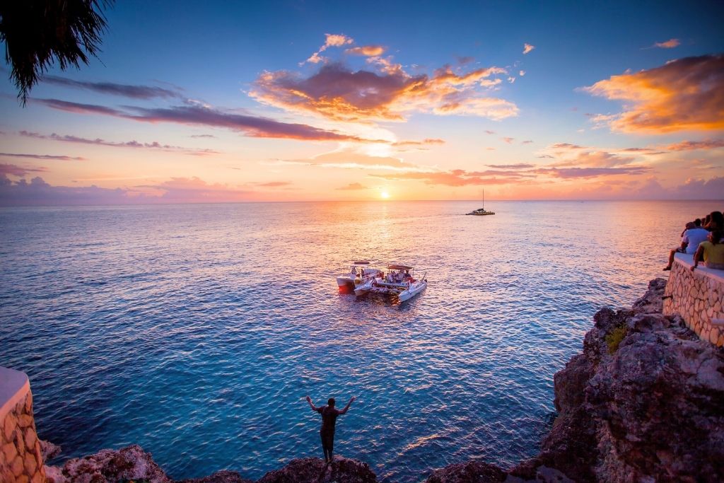 A glowing sunset over the ocean in Negril, Jamaica, with boats floating near the cliffs and a silhouetted figure raising their arms in celebration at the edge.