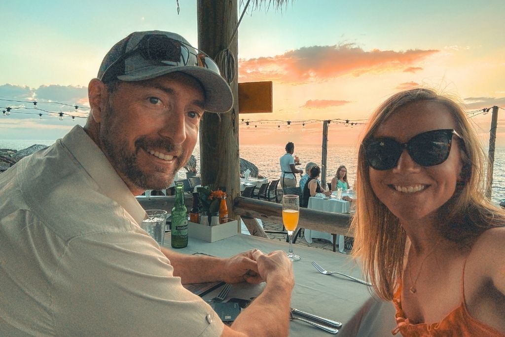 Kate and her husband smile at a seaside restaurant table in Jamaica during sunset, with drinks, string lights, and a glowing ocean backdrop behind them.