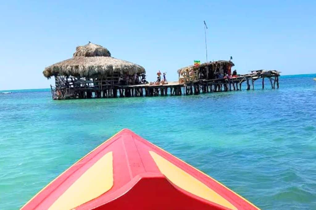 A colorful boat approaches the thatched-roof Pelican Bar, a rustic wooden bar built on stilts over the turquoise sea off the coast of Jamaica.