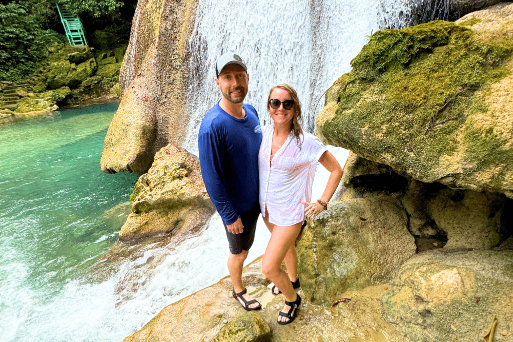 Kate and a man smile together beside a lush waterfall in Jamaica, standing on mossy rocks with clear blue water cascading behind them.