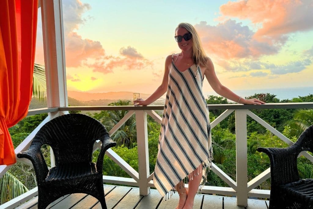 Kate stands on a balcony at sunset in Jamaica, wearing a striped sundress and sunglasses. Lush greenery and colorful clouds glow in the sky behind her.
