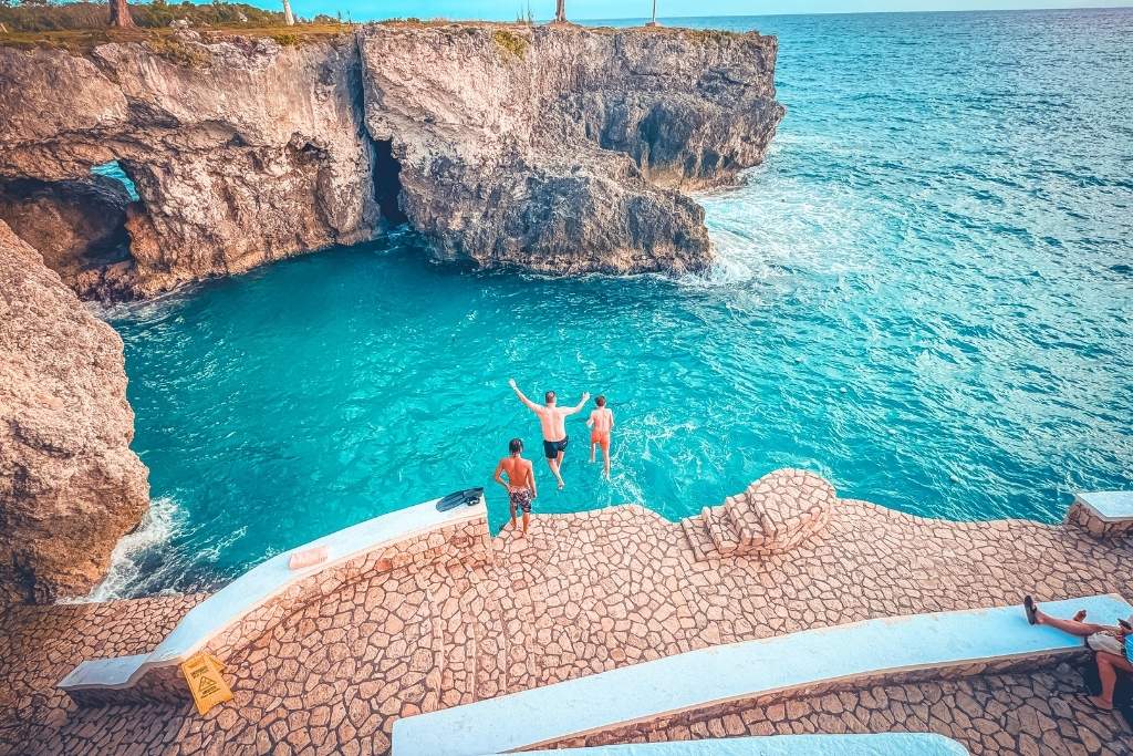 Two people leap into bright turquoise water from a stone terrace at Rick’s Cafe in Negril, Jamaica, with dramatic cliffs and ocean views all around.