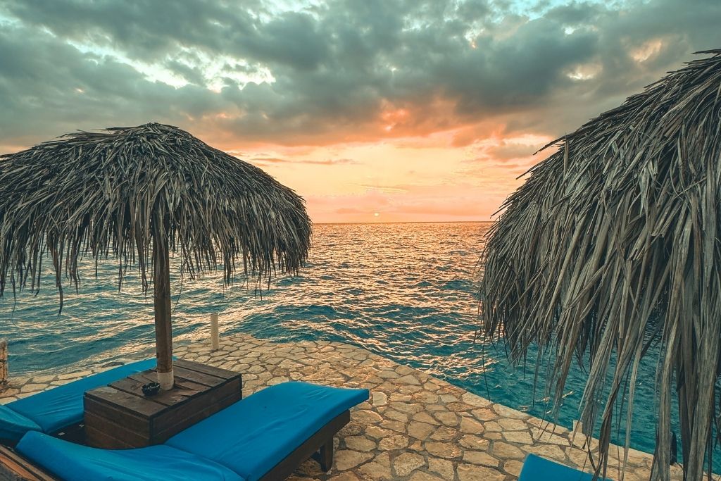 A sunset view over the ocean in Jamaica with two lounge chairs under thatched umbrellas on a stone terrace, capturing a calm and dreamy tropical evening.