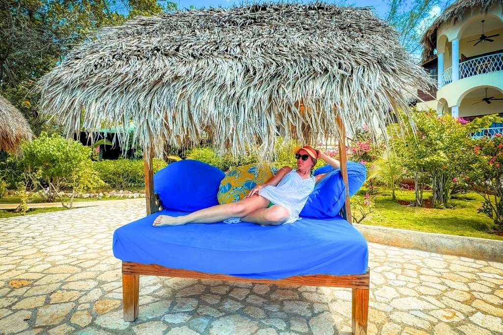 Kate lounges under a thatched cabana on a bright blue cushioned daybed at a tropical resort in Jamaica, surrounded by lush gardens and sunshine.