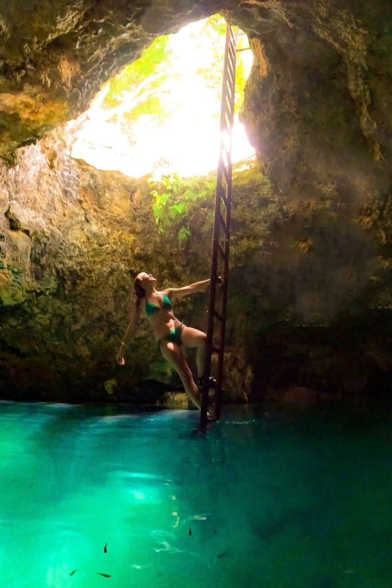 Kate climbs a rusty ladder from glowing turquoise water inside the Blue Hole Mineral Spring cave, with sunlight streaming in from the rocky opening above.