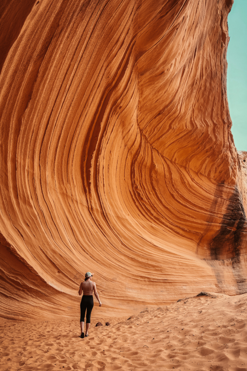 Kate walks along the sandy floor of a towering sandstone wave formation near Page, Arizona, with striated red rock curving dramatically above her under a pale sky.