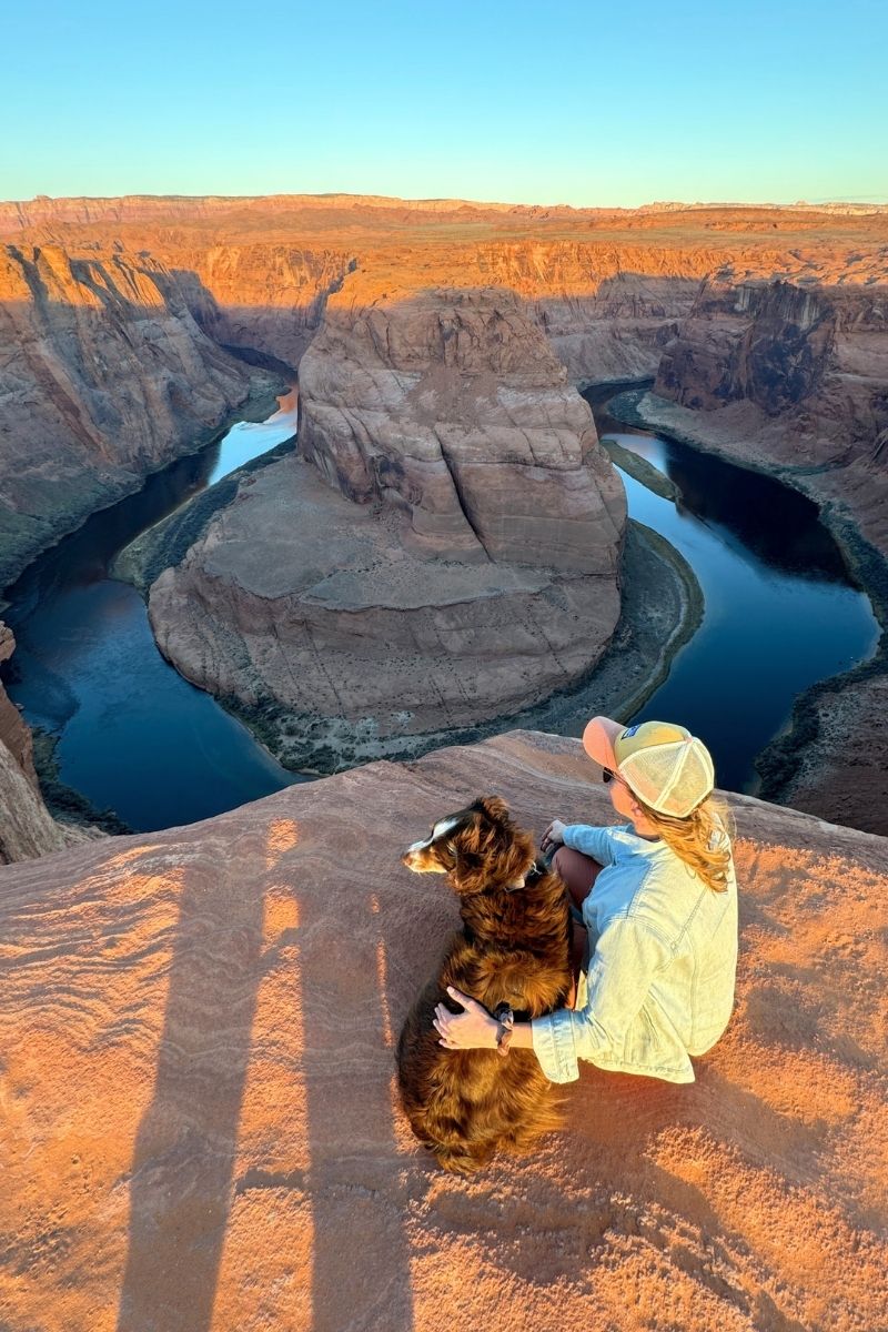 Kate sits with her dog at the edge of Horseshoe Bend near Page, Arizona, both looking out over the dramatic curve of the Colorado River and red rock canyon at sunset.