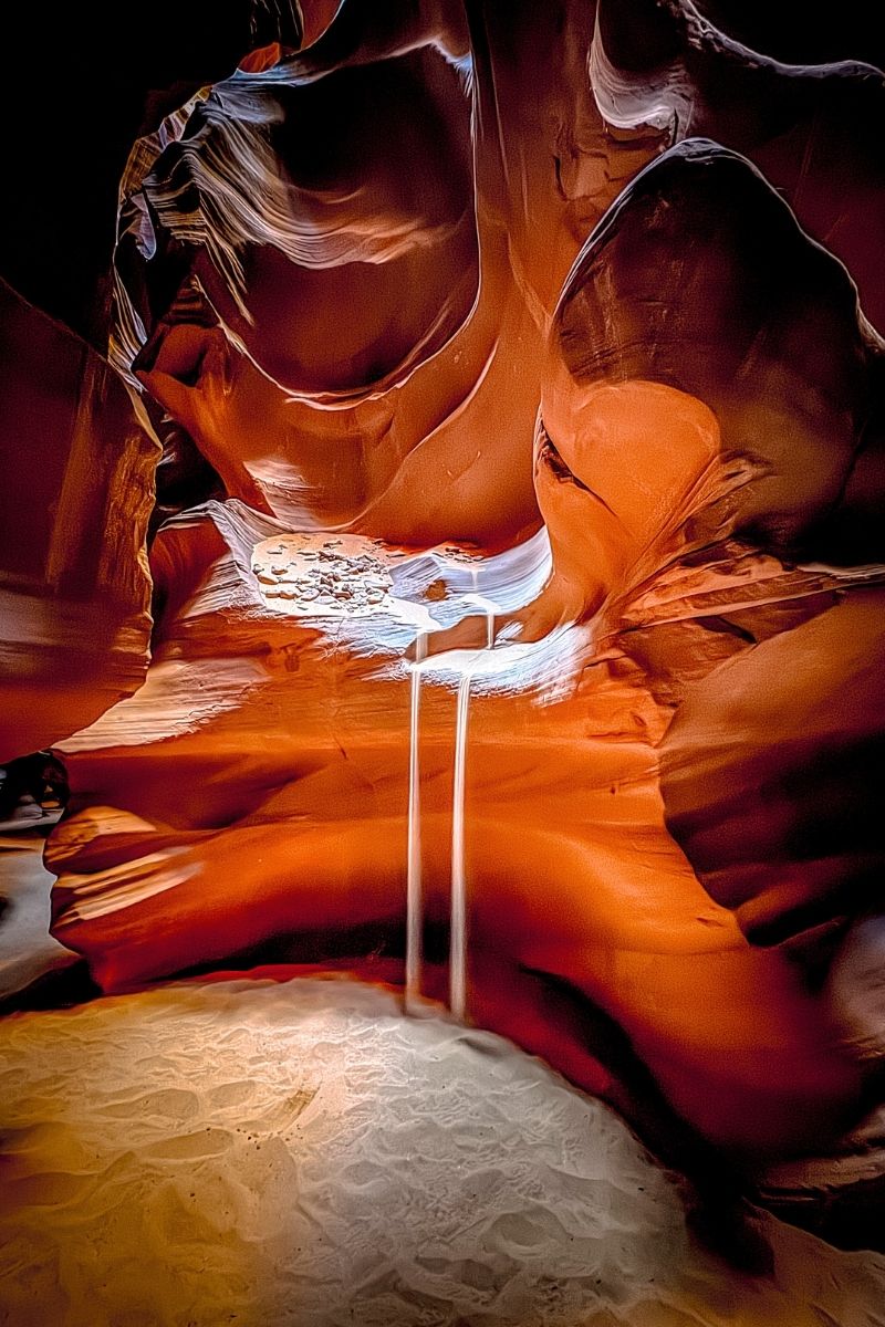 Fine sand pours like a waterfall from a ledge inside Upper Antelope Canyon, framed by smooth, glowing red sandstone walls and a patch of sunlight illuminating the canyon floor.