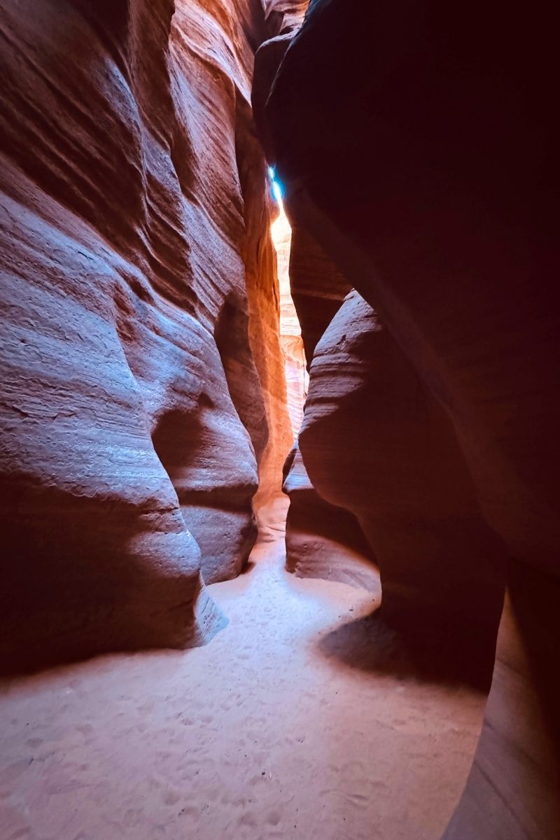 A narrow path winds through the softly lit curves of Canyon X near Page, Arizona, with smooth red and purple sandstone walls towering on both sides and light filtering from above.