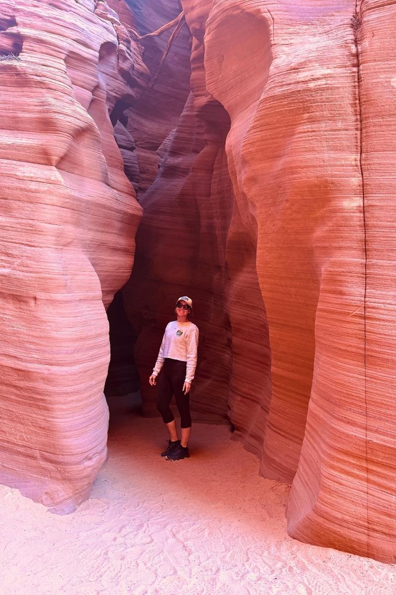 Kate stands smiling inside the vibrant pink and orange Canyon X near Page, Arizona, surrounded by smooth, striated sandstone walls that curve gracefully around her.