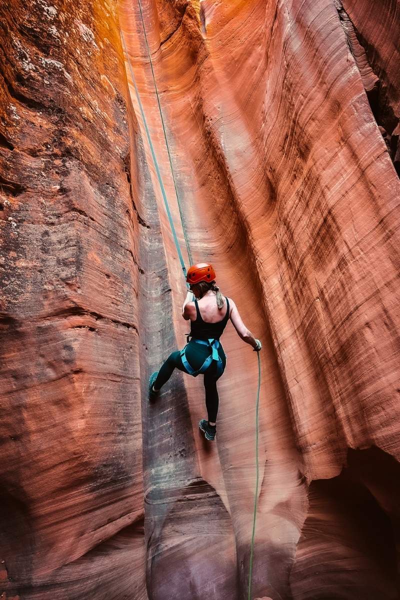 Kate rappels down a narrow red rock wall at Antelope Ridge Adventure Park near Page, Arizona, wearing a helmet and harness as she descends through a colorful slot canyon.