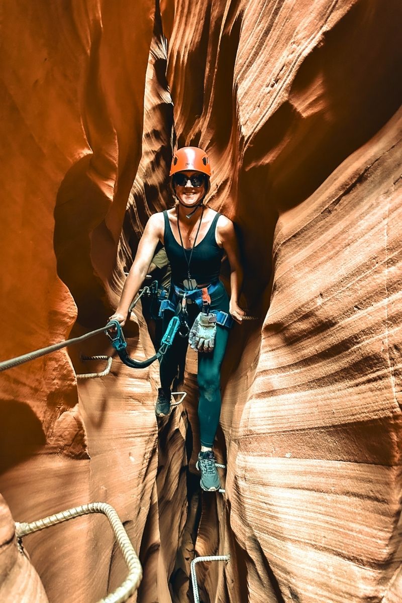 Kate smiles while climbing through a narrow sandstone slot canyon at Antelope Ridge Adventure Park, using a harness and metal rungs secured to the rock walls for support.
