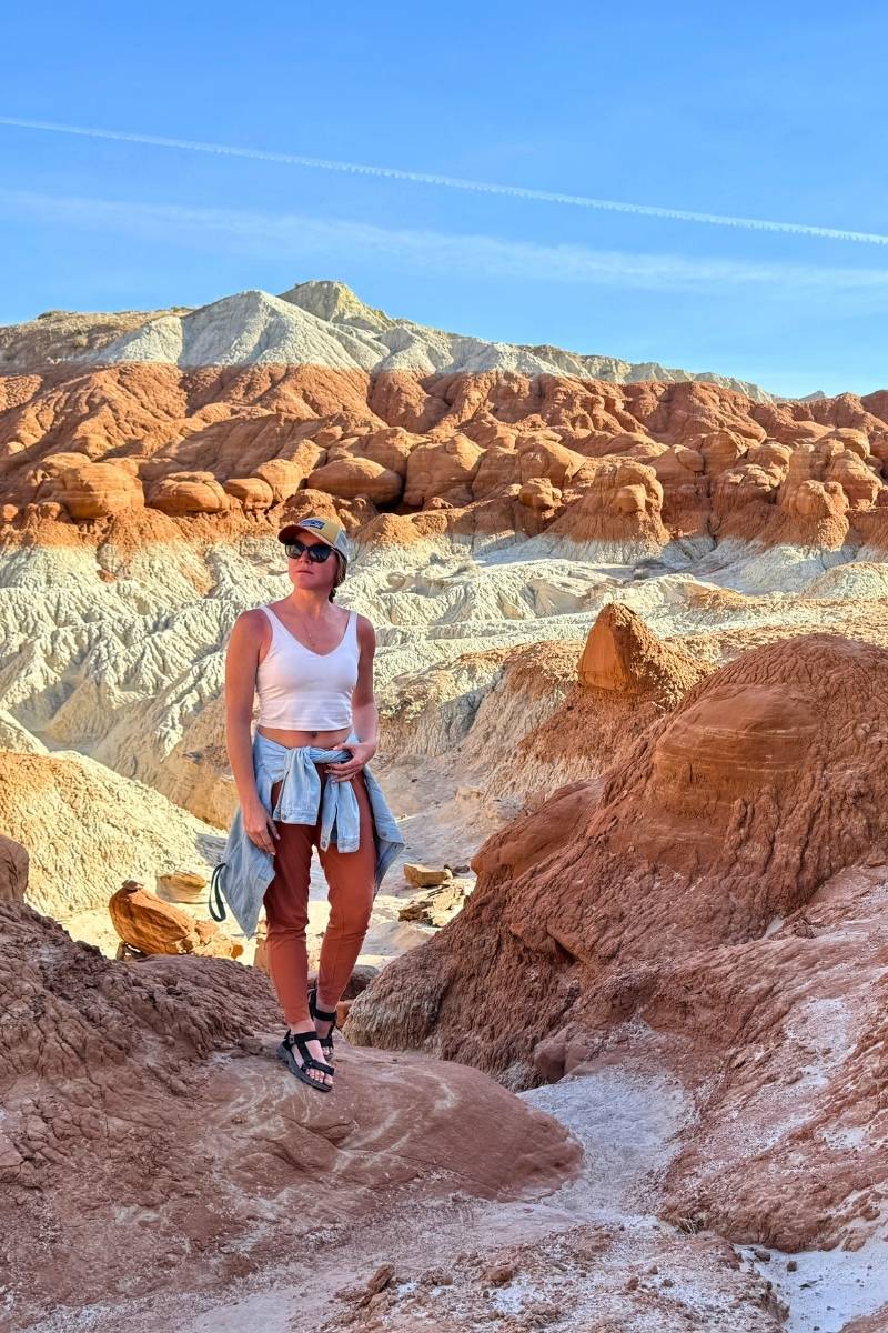 Kate stands along the Toadstool Hoodoos Trail near Page, Arizona, surrounded by layered red and white rock formations under a clear blue sky, dressed for a desert hike.
