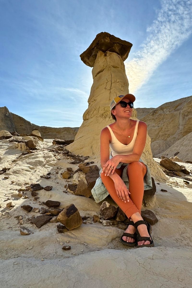 Kate sits smiling at the base of a tall white hoodoo on the Toadstool Hoodoos Trail near Page, Arizona, surrounded by scattered rocks and sunlit desert formations.