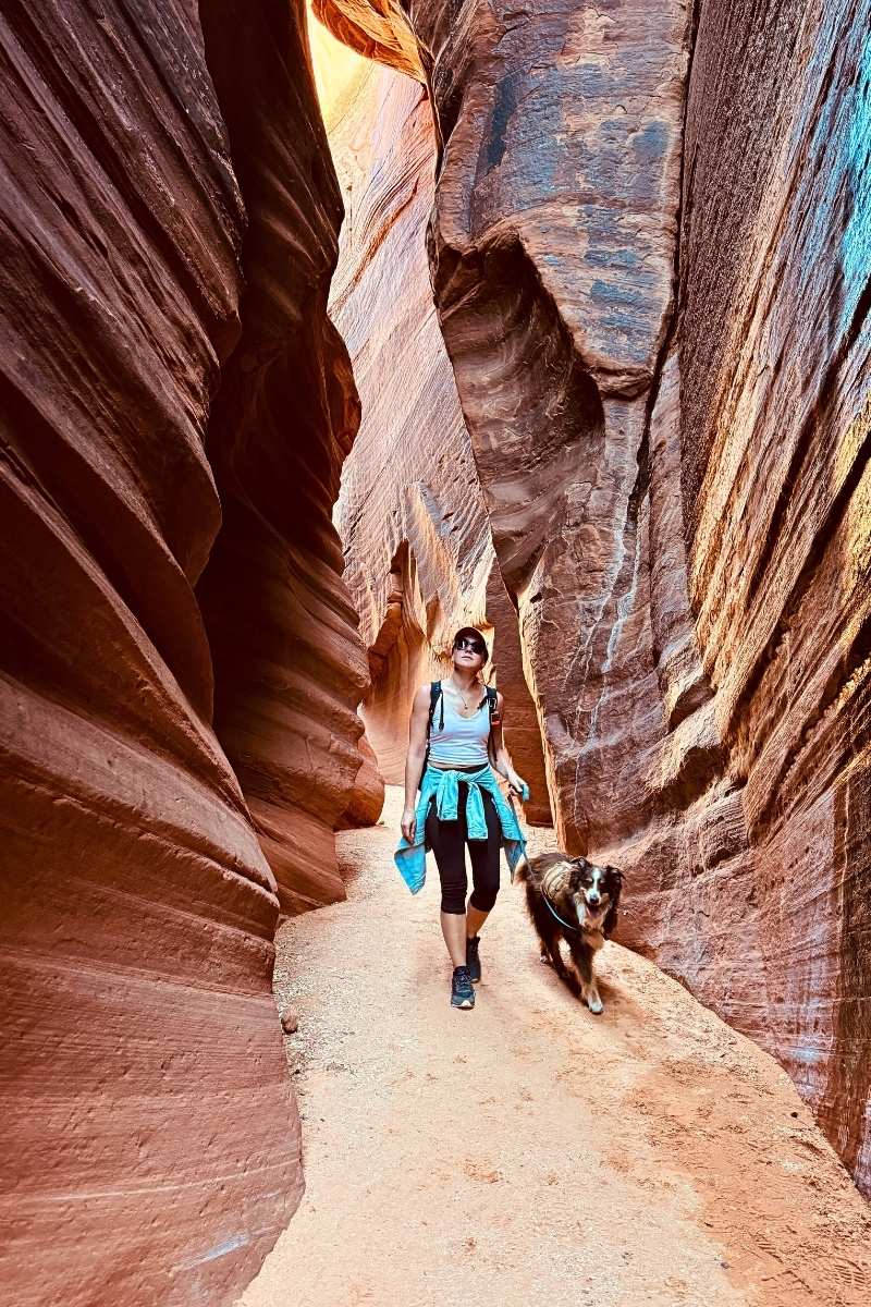 Kate hikes through a narrow red rock slot canyon on Wire Pass Trail with her dog by her side, surrounded by tall, curving sandstone walls in warm desert light.