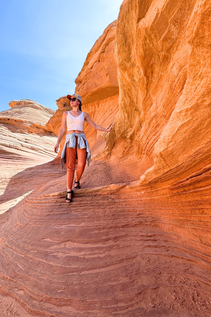 Kate walks confidently along smooth, curving red rock formations on the Beehive Trail near Page, Arizona, with one hand brushing the sandstone and sunlight highlighting the natural textures.