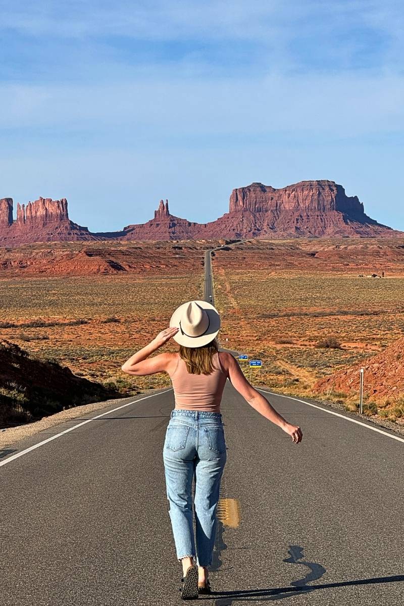Kate wearing a wide-brimmed hat and jeans walks down the iconic straight road toward Monument Valley’s red rock formations, framed by a clear blue sky and vast desert landscape.