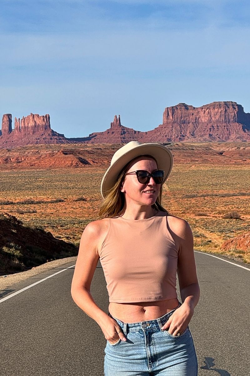 Kate in a tan crop top, jeans, and a wide-brimmed hat stands smiling on a desert highway with Monument Valley’s iconic red rock buttes rising behind her under a clear blue sky.