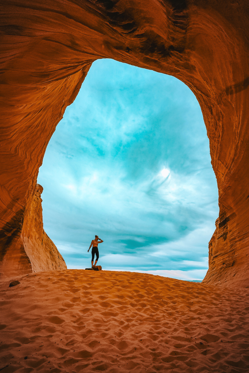 Kate stands at the edge of a sunlit sand dune inside a dramatic red rock cave near Page, Arizona, looking out toward the bright blue sky through the arching rock opening.