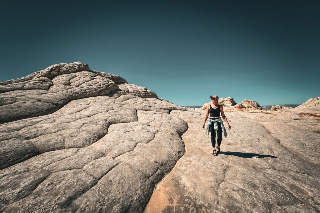 Kate hikes across a cracked, moonlike rock formation under a deep blue sky in the desert near Page, Arizona, wearing a cap, tank top, and leggings with a jacket tied around her waist.