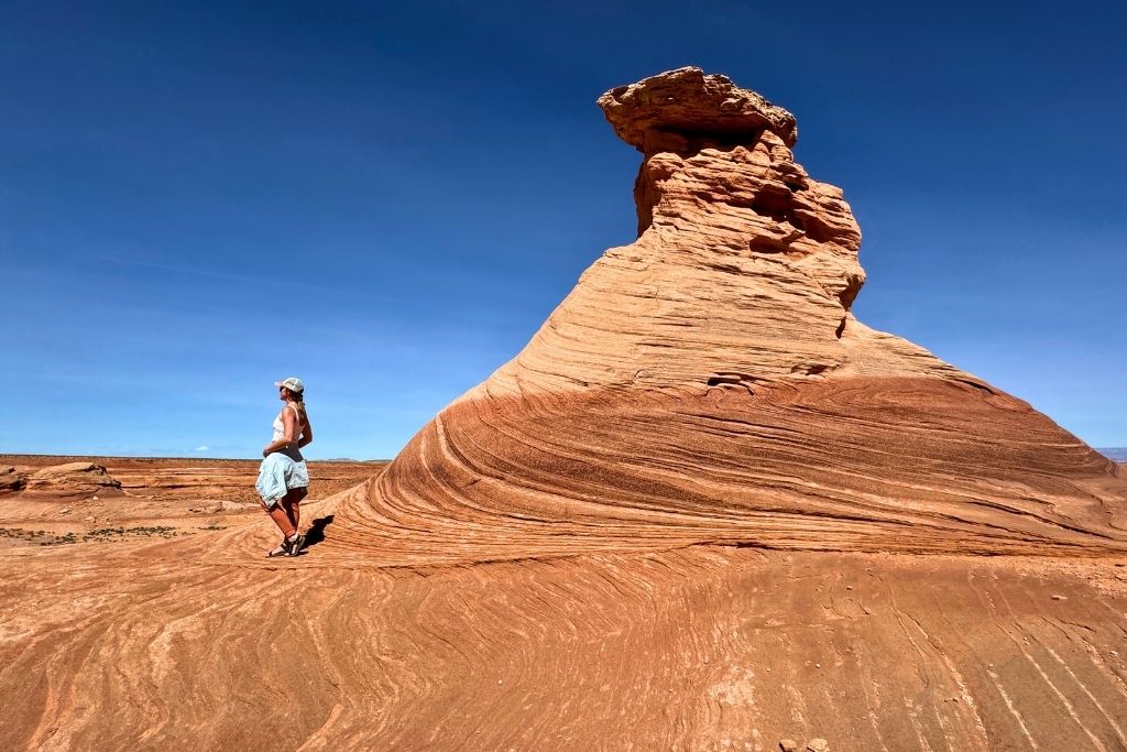 Kate stands beside a swirling, layered rock formation known as the Beehive on a sunny day in Page, Arizona, with vivid red sandstone and a clear blue sky in the background.
