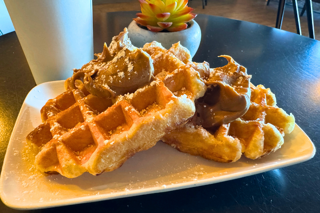 A plate of golden, fluffy waffles topped with generous scoops of cookie dough butter and a dusting of powdered sugar, served at Lake Powell Espresso with a white coffee cup and a small succulent plant in the background.