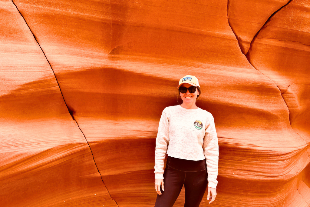 Kate standing and smiling in front of a smooth, vibrant red-orange sandstone wall with natural striations and cracks, typical of the rock formations found in Page, Arizona. She’s wearing a light sweatshirt, black leggings, sunglasses, and a hat.