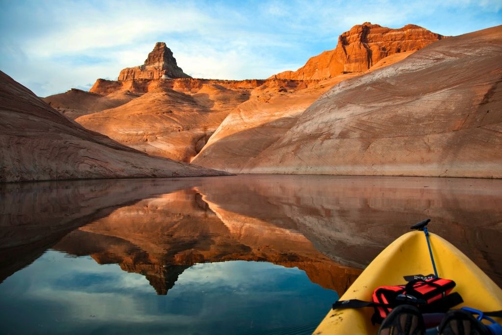 A peaceful view from a yellow kayak on Lake Powell, with calm water perfectly reflecting the surrounding red rock formations and distant peaks glowing in the golden light.