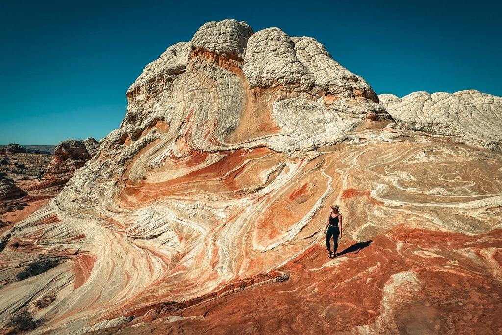 Kate walks across the swirling, multicolored rock landscape of White Pocket in Vermilion Cliffs National Monument, surrounded by dramatic white and red sandstone formations under a clear blue sky.