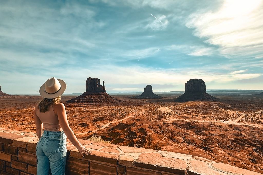 Kate in a wide-brimmed hat and jeans stands at a scenic overlook in Monument Valley, gazing out at the towering buttes and vast red desert landscape under a dramatic sky.